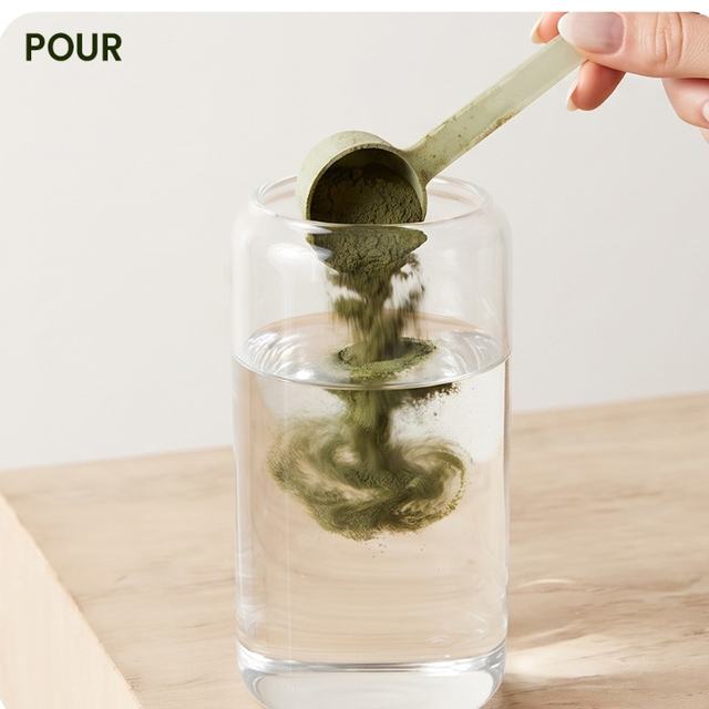 A scoop of green powder falling into a glass of cold water